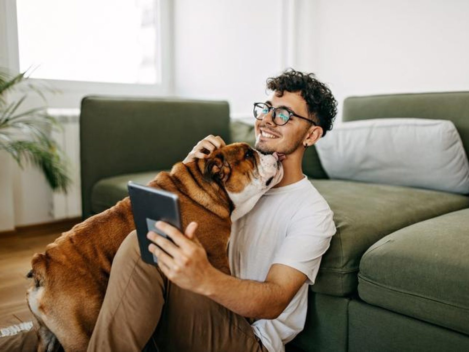 A person holds a tablet displaying a real estate app while sitting on the floor with their pet dog.