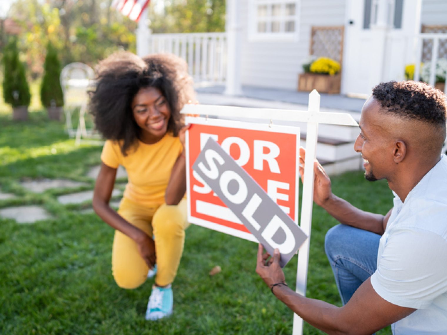 A joyful man and woman are interacting near a "FOR SALE" sign in front of a house. The woman is crouching down and smiling broadly while the man, in a crouched position as well, is attaching a "SOLD" sticker to the sign.