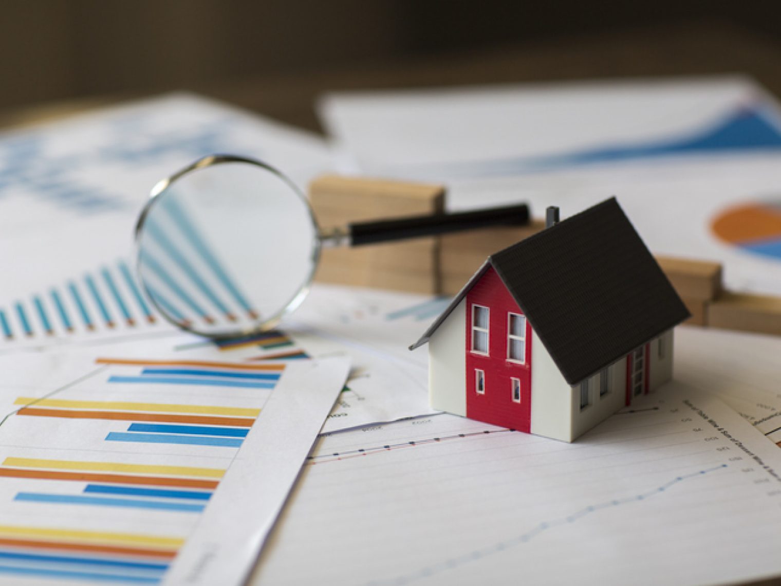 A small model house sits on top of financial charts and graphs with a magnifying glass.