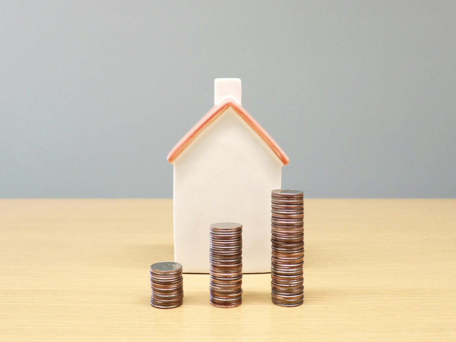 Small white house figurine behind three ascending stacks of coins on a wooden table.