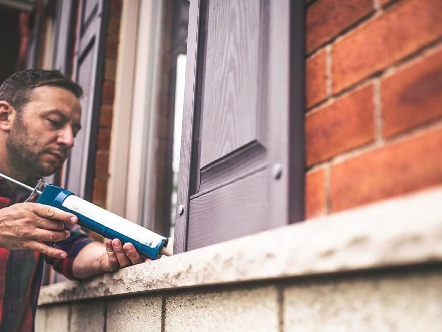 A man applies caulk to the window of a brick home.