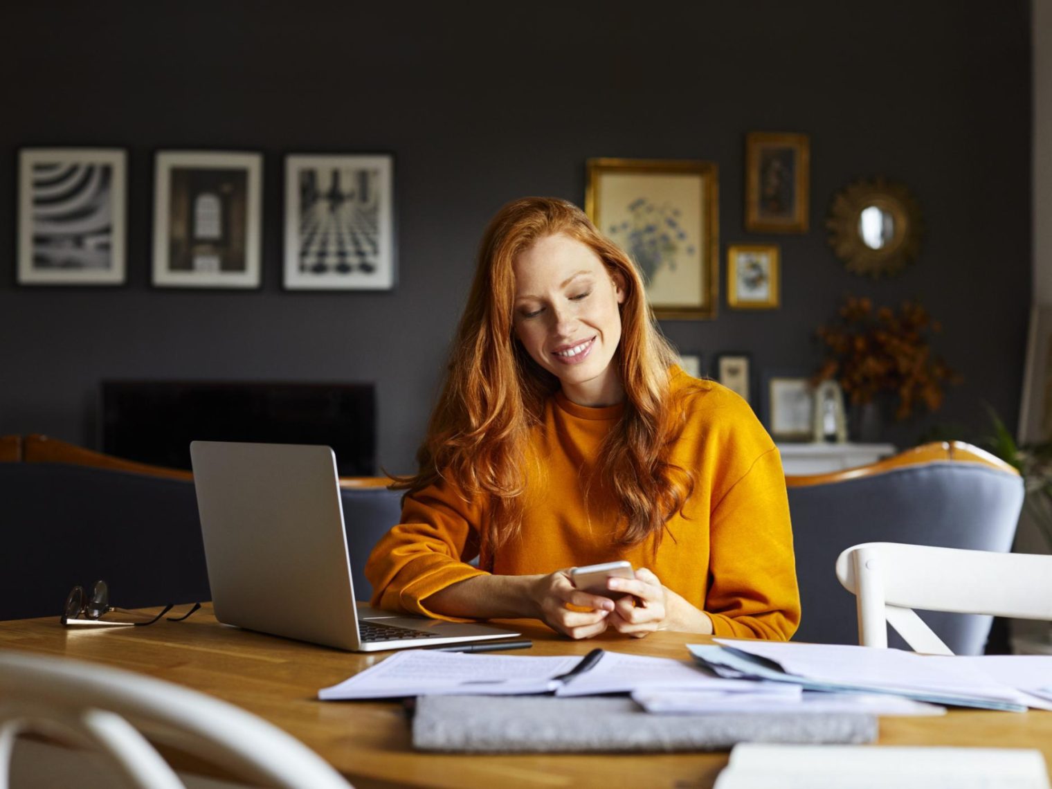 A smiling woman sitting at a table in her home with a laptop and mobile phone.