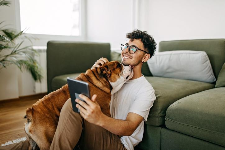 A person holds a tablet displaying a real estate app while sitting on the floor with their pet dog.
