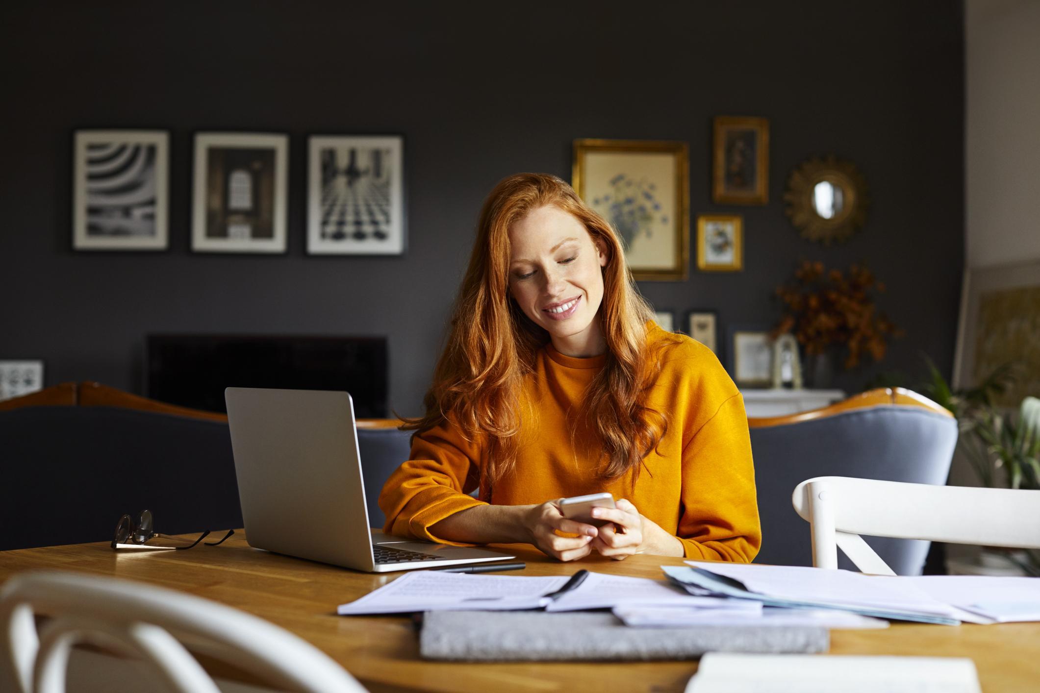 A smiling woman sitting at a table in her home with a laptop and mobile phone.